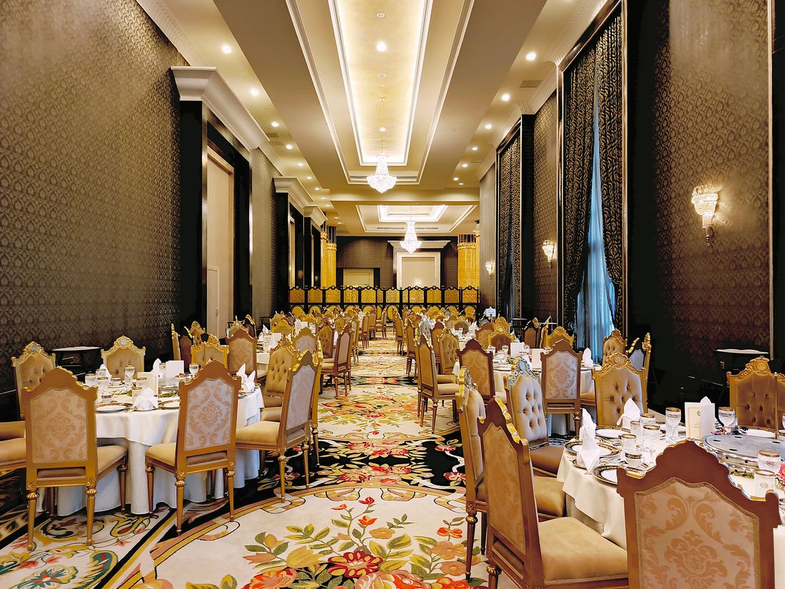 An opulent empty ballroom set for a formal dinner, round tables with white linens and gold-upholstered chairs under warm chandelier light.