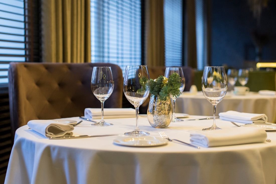 A round dinner table set with white linens, folded napkins, wine glasses, and a small evergreen centerpiece catching warm ambient light, no people in frame.