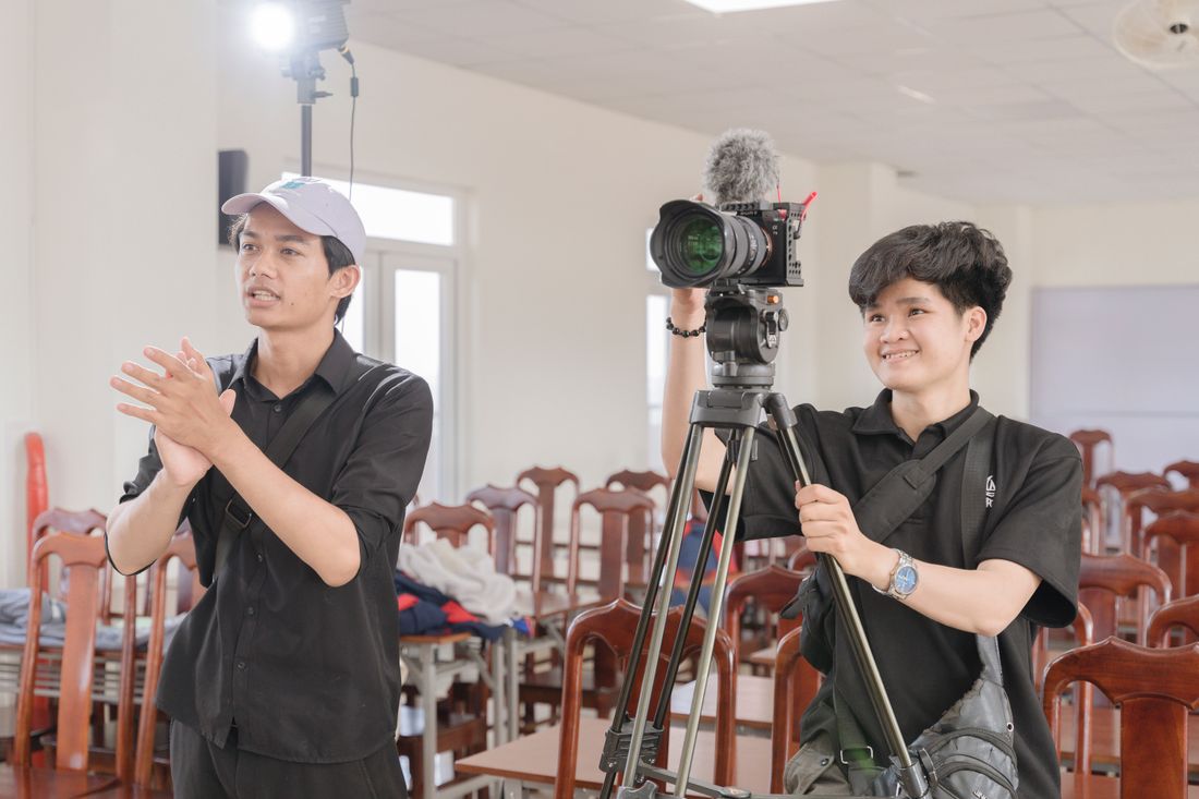 Two video crew members setting up a camera on a tripod in a bright, plain room with rows of wooden chairs and a whiteboard behind them.