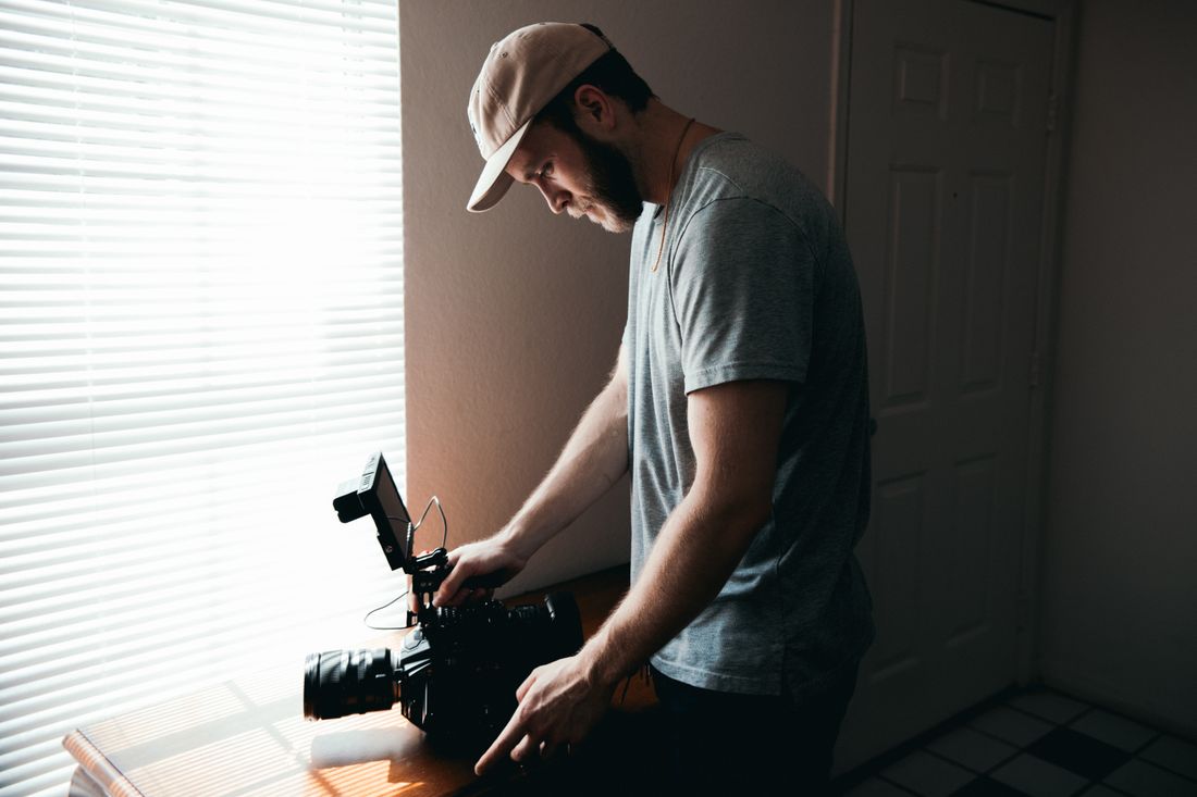A videographer in a ball cap adjusting a professional video camera on a table beside a sunlit window with blinds.