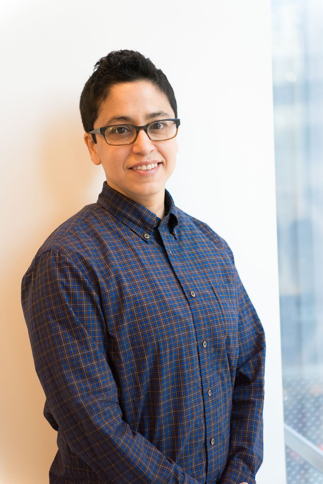 A friendly professional in a casual plaid shirt and glasses poses for a portrait indoors near a window.