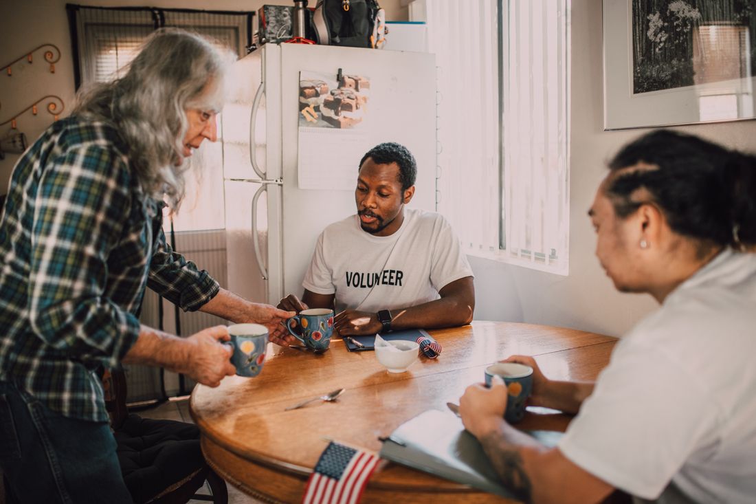 A small group shares coffee and conversation around a kitchen table in a modest community setting.