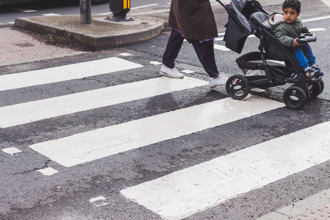 A tight crosswalk view of a parent pushing a child in a stroller across a street crossing.