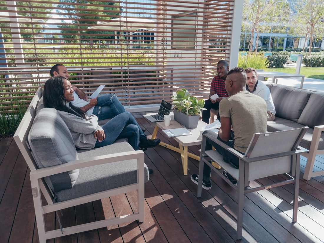 Two small clusters of colleagues talk on a covered outdoor deck, some seated on lounge furniture, in a relaxed daytime setting.