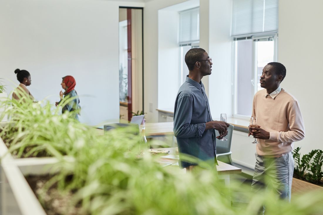 Two colleagues hold champagne glasses and talk in a bright office with large windows, plants in the foreground and other coworkers chatting in the background.