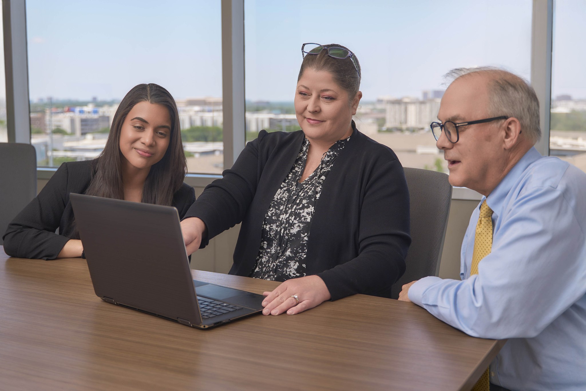 Three colleagues look at an open laptop together at a conference table, with a city skyline visible through large windows.