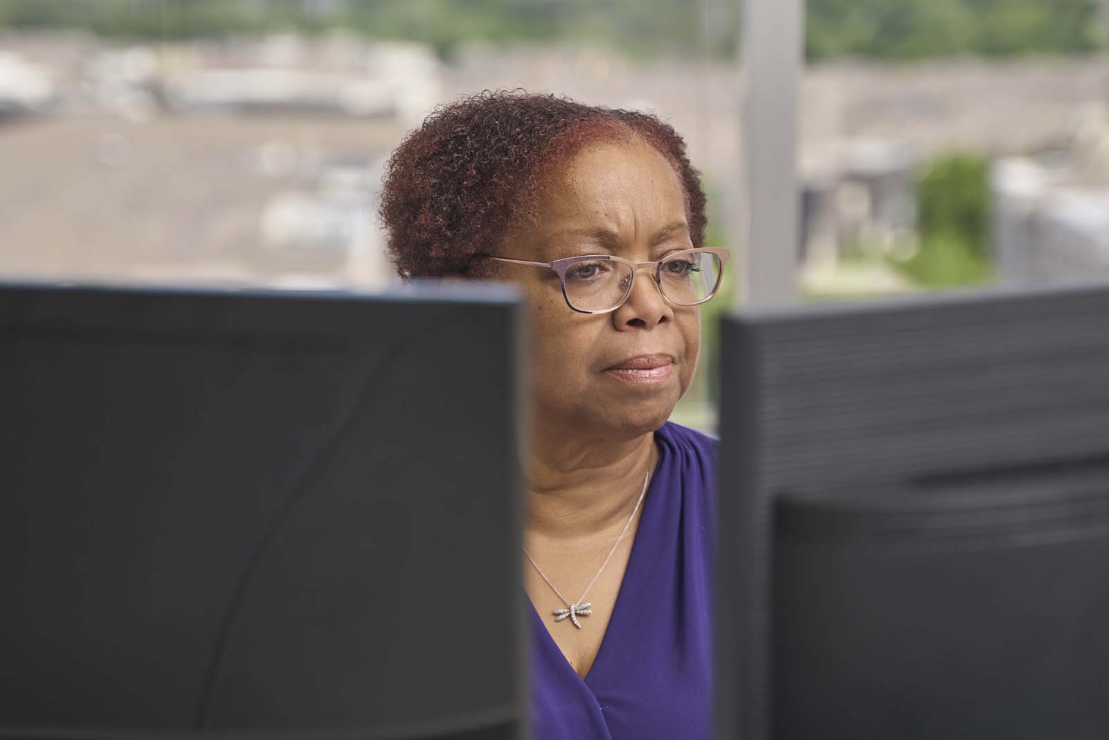 An older woman in a purple top studying a monitor, seen through the gap between two cubicle partitions, the kind of candid focused-work moment that reads as authentic rather than staged.
