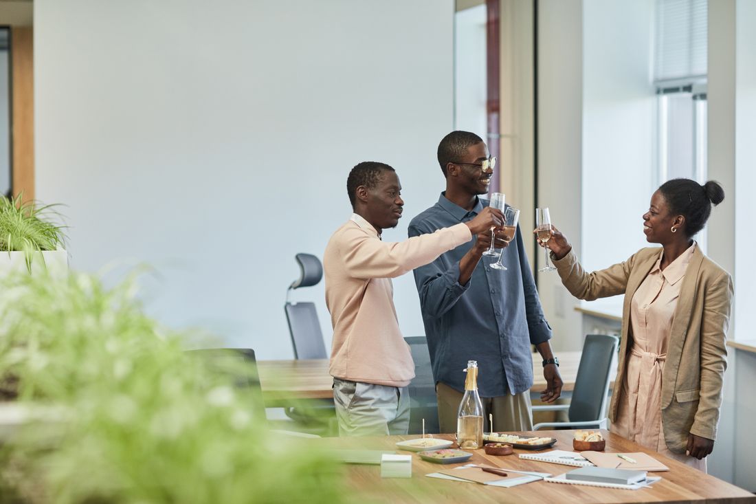 Three colleagues in business-casual clothing stand around a table in a bright office common area, raising glasses in a toast.