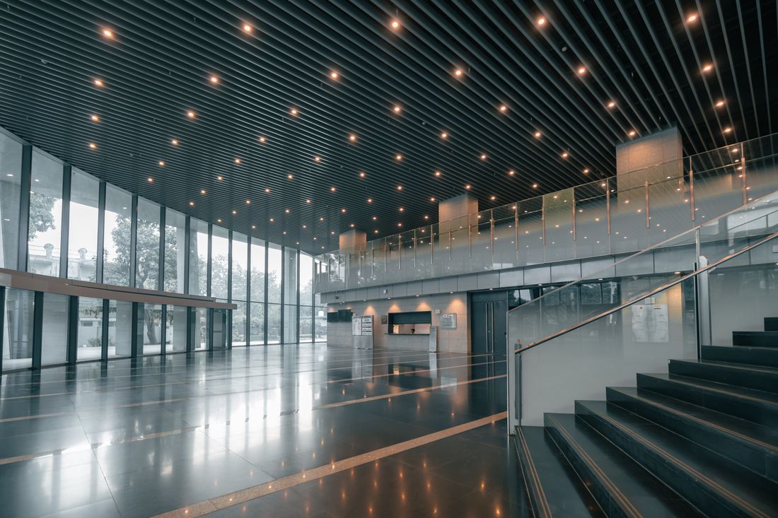A large, empty modern building lobby with a glass curtain wall, polished stone floor, and an open staircase rising to a mezzanine.