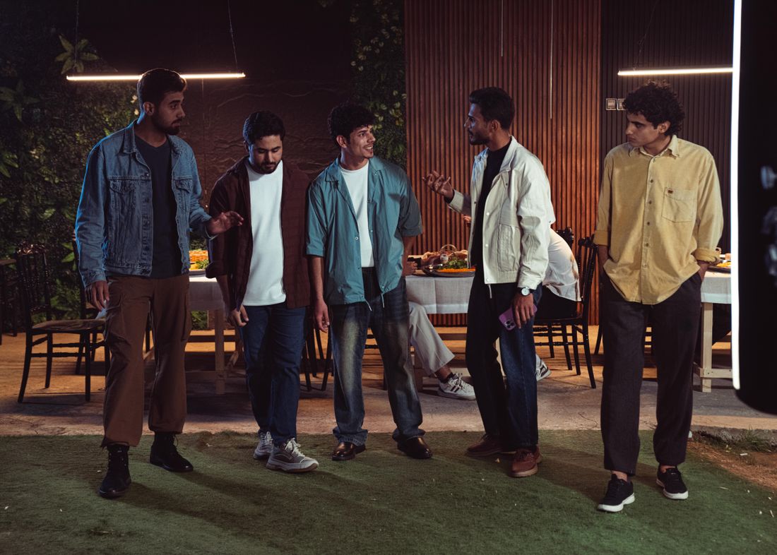 A group of colleagues in casual clothing chat and laugh around an outdoor dinner table lit by string lights in the evening.