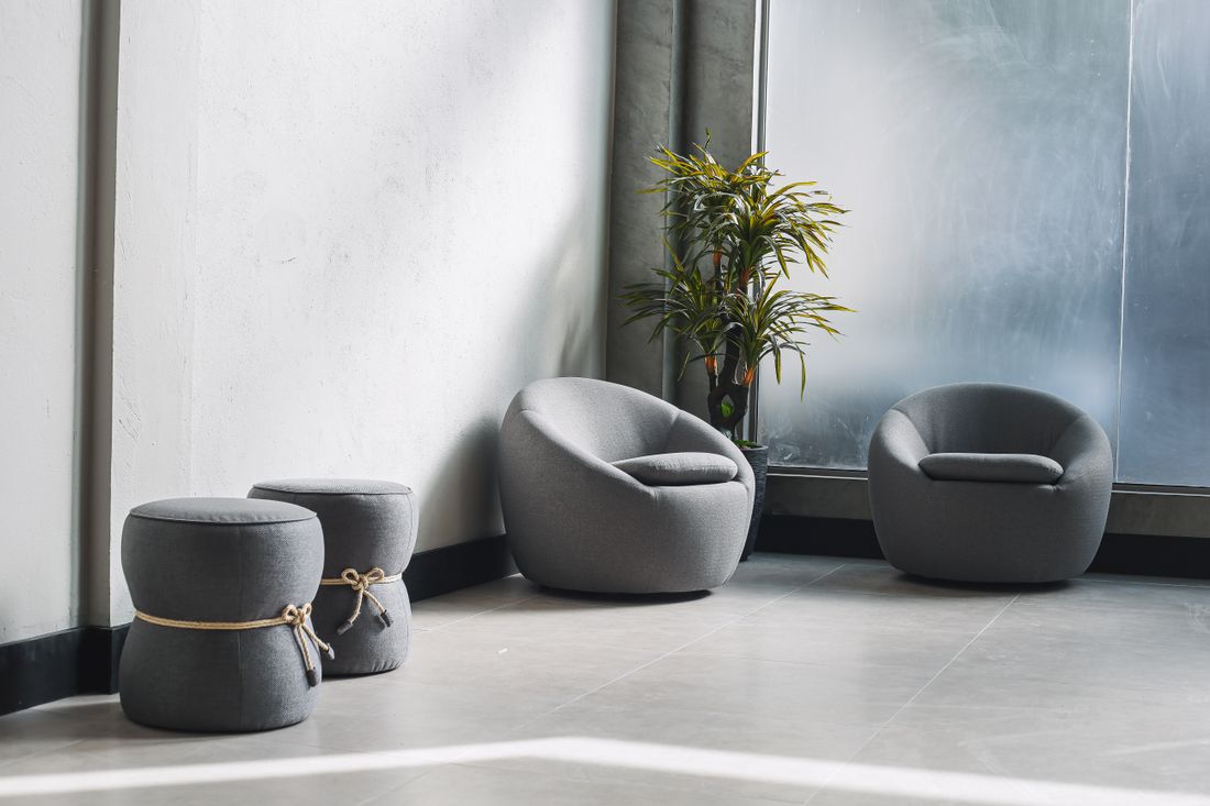An empty modern lounge with soft gray armchairs, tied ottomans, and a potted plant beside a bright window.