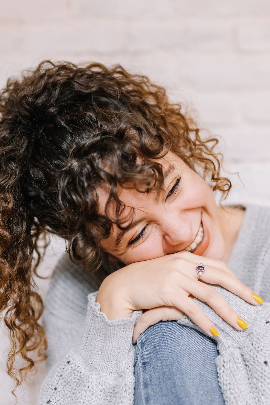 A woman with curly hair laughs warmly, resting her chin on her knee, against a plain blurred background.
