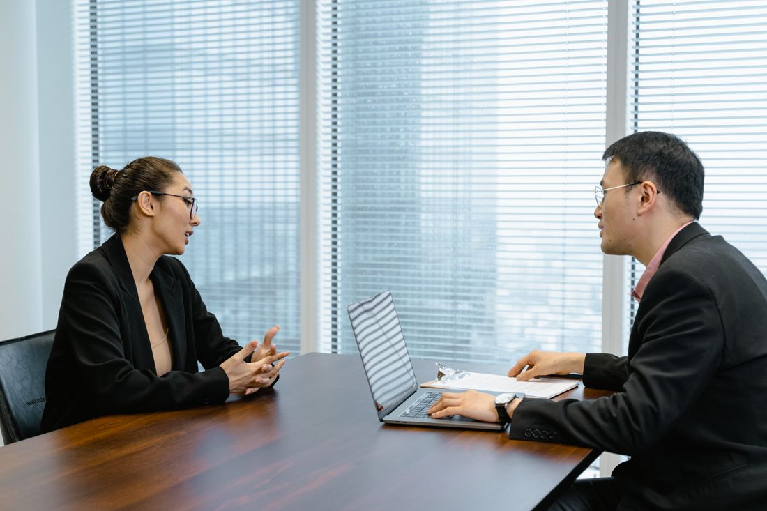 Two professionals in business attire in conversation across a table in a modern office, floor-to-ceiling windows behind them
