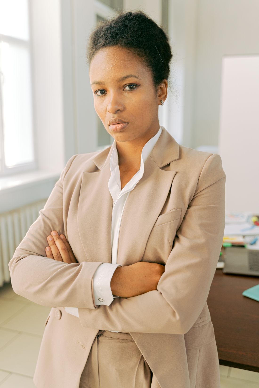 Confident professional woman in a tan blazer standing with arms crossed, softly blurred office background