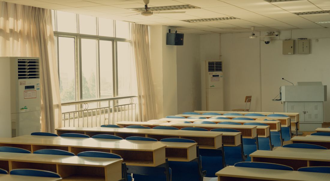 An empty training room with rows of desks and blue chairs facing a podium, morning light coming through tall windows.