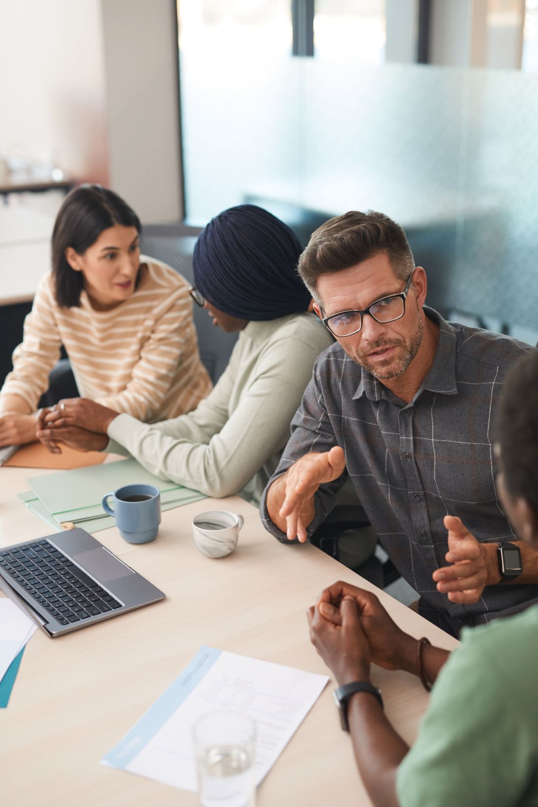 A small group of colleagues talk around a table with a laptop, coffee cups, and papers in front of them.