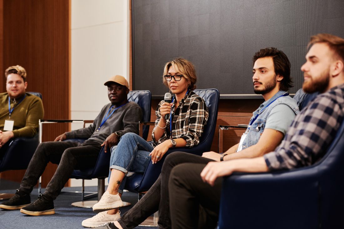 Five panelists seated in chairs on a conference stage, one speaking into a handheld microphone while the others listen.