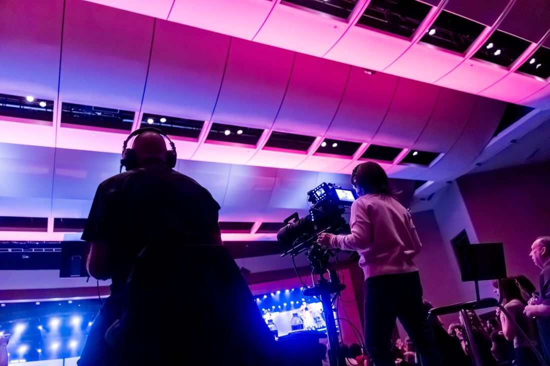 Two video camera operators filming a live event from the back of a room under colored stage lighting.