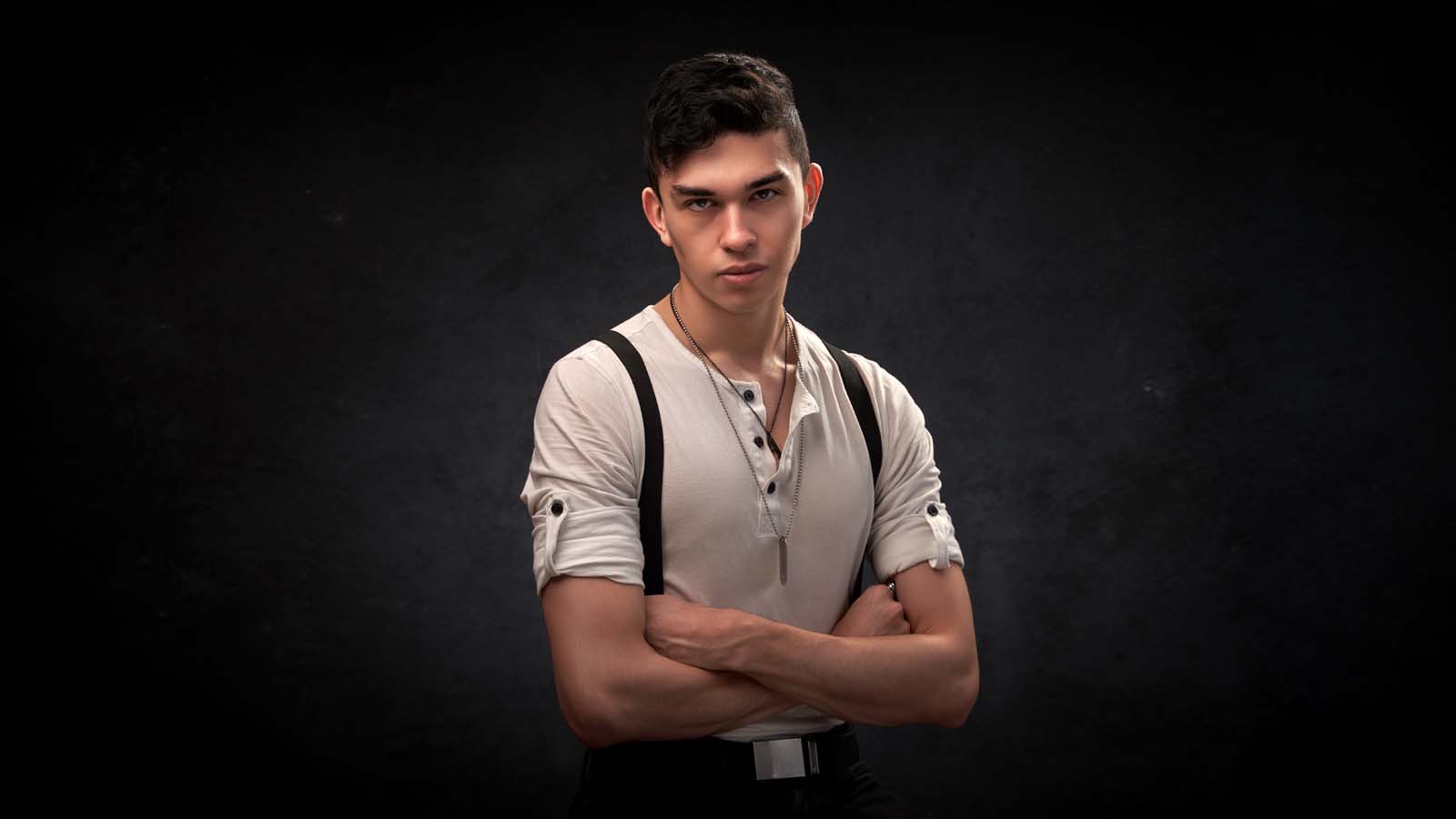 Young man with dark hair in a white henley and dark suspenders, arms crossed, against a black studio backdrop