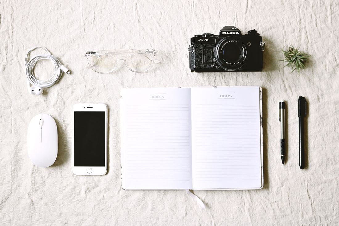 Blank open notebook labeled 'notes' laid out with a camera, phone, glasses, and headphones on a flat surface