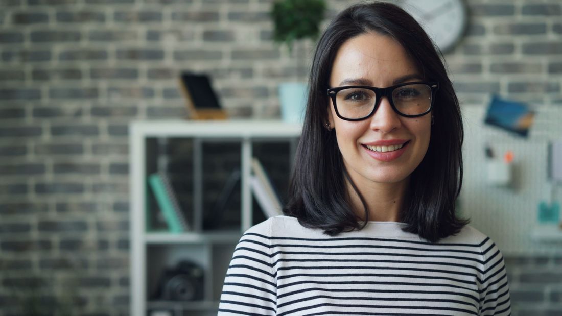 Consultant in glasses smiling in a real office workspace with a brick wall background, an approachable professional portrait