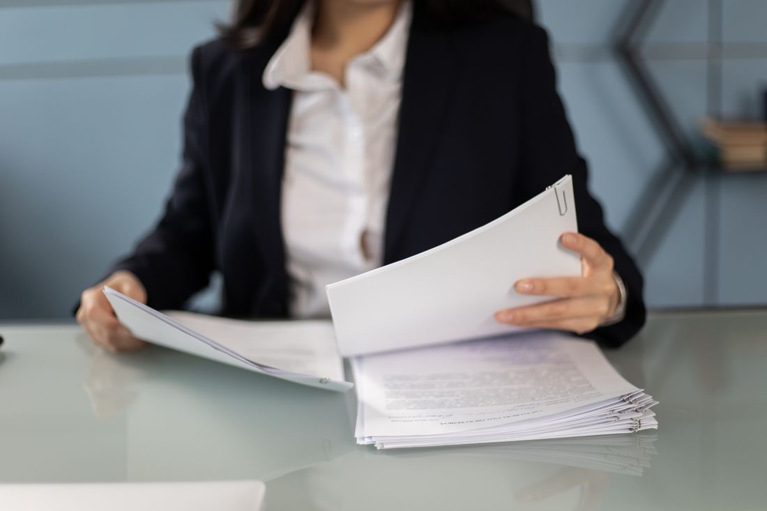 Over-the-shoulder view of a person in a blazer comparing two paperclipped stacks of printed documents on a glass desk.