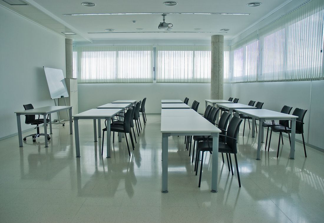 Empty corporate training room set up before a session, with rows of tables and chairs, a blank flip chart, and daylight through vertical blinds.