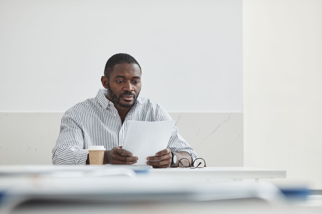 A person seated at a desk reading a blank sheet of paper, with a coffee cup and glasses nearby.