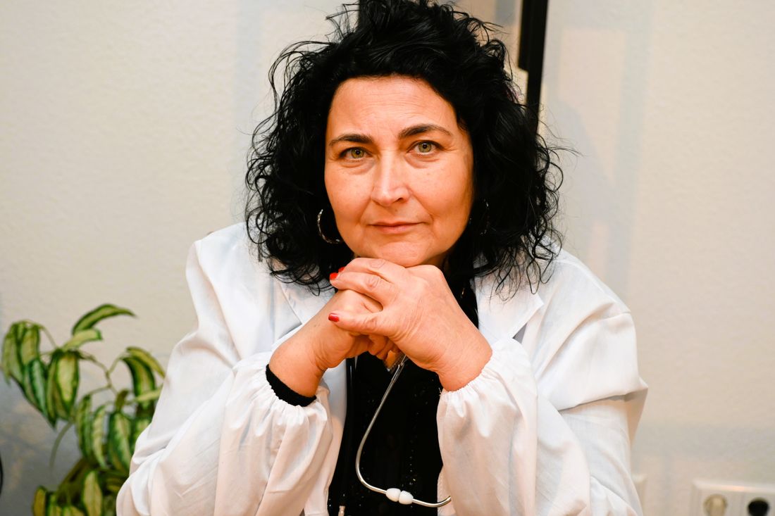 A woman in a white coat sits for a relaxed portrait, stethoscope draped around her neck, hands folded, looking directly at the camera against a plain wall.