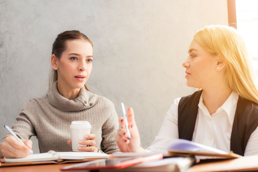 Two women in business-casual clothing sit at a small table talking, one gesturing mid-sentence with a pen and coffee cup in hand, the other listening.