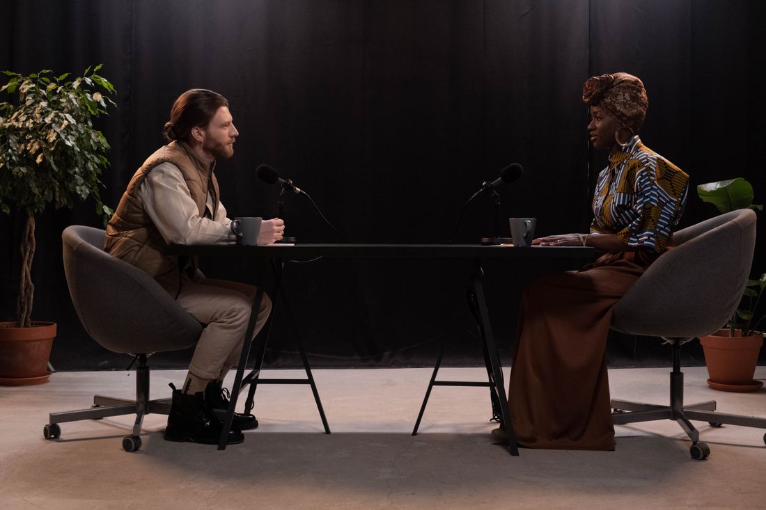 A guest and a host seated across a table from each other during a podcast recording, each speaking into a microphone.