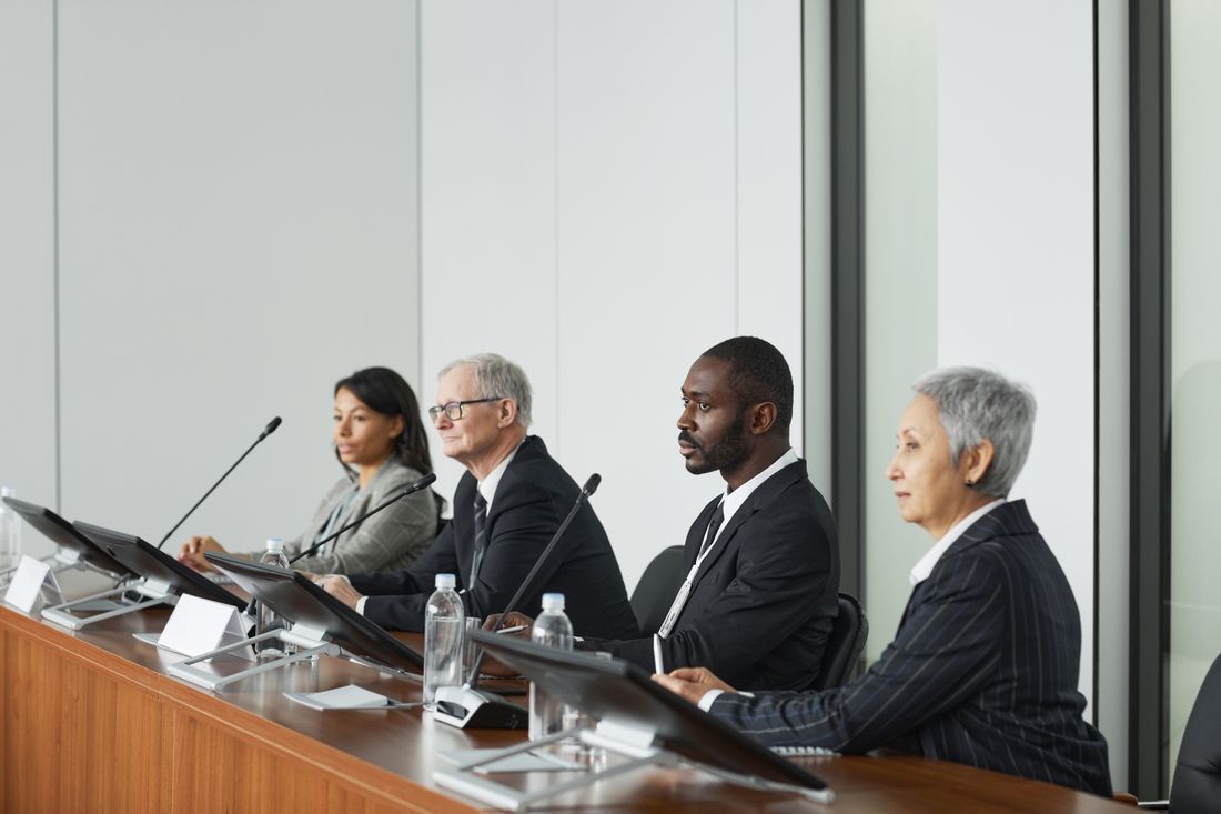 Four panelists seated in a row at a conference table, each with a microphone and name placard, during a panel discussion.