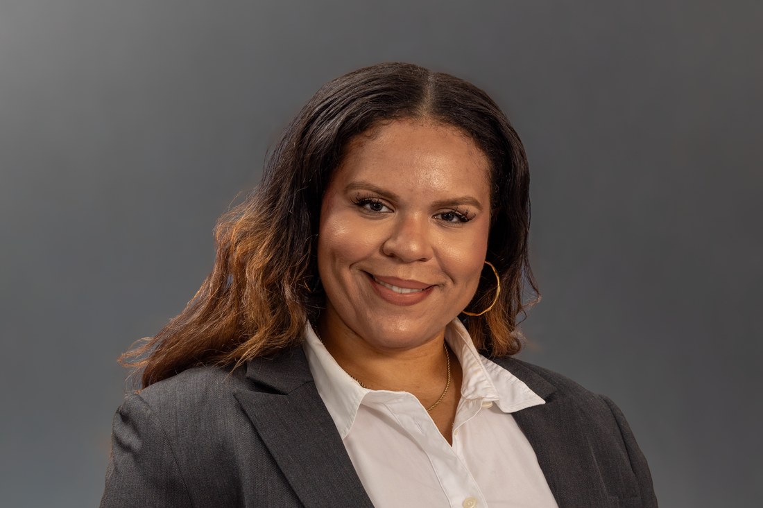 Studio portrait of a smiling woman in a charcoal blazer and white collared shirt against a plain grey background.