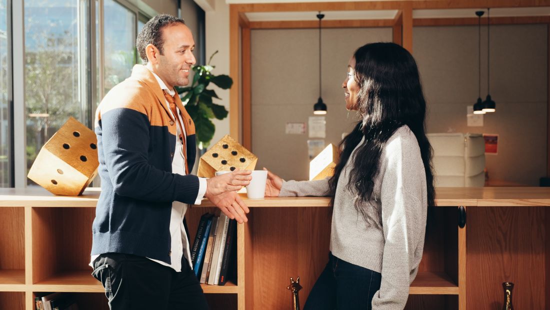 Two colleagues in business-casual sweaters standing and talking over coffee at a wood counter in a bright, plant-filled office.