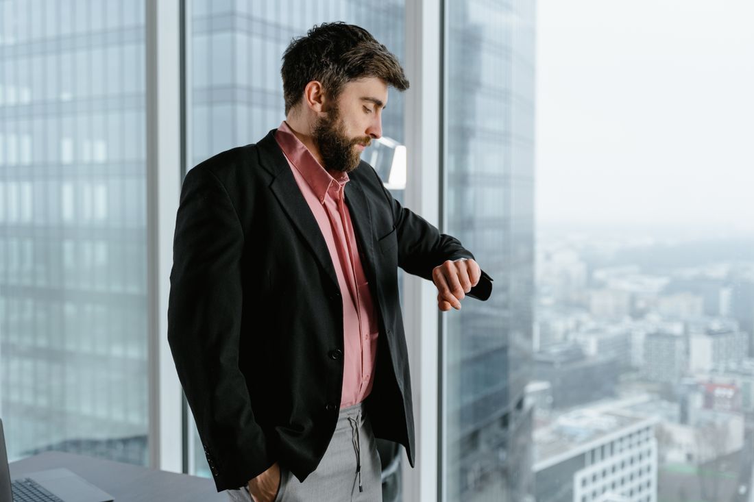 A man in a dark suit stands in profile checking his watch beside a floor-to-ceiling office window with a hazy city skyline in the background.