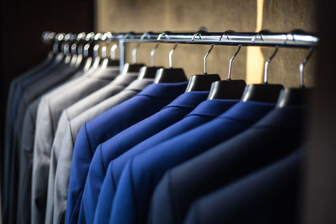 A row of plain gray and navy suit jackets hangs neatly on a metal rail against a warm neutral wall, with no people in frame.