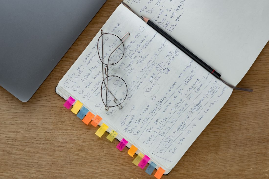 A pair of eyeglasses rests on an open notebook with handwritten notes and a pen on a wooden desk.