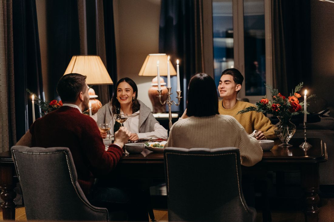A small group of adults seated around a candlelit dinner table in a warm private room, talking and smiling with wine glasses and a silver candelabra in view.
