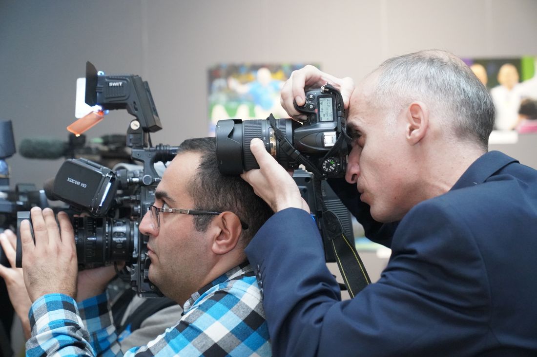 Two photographers with professional cameras and a shoulder-mounted video rig shoot side by side indoors at a press event.