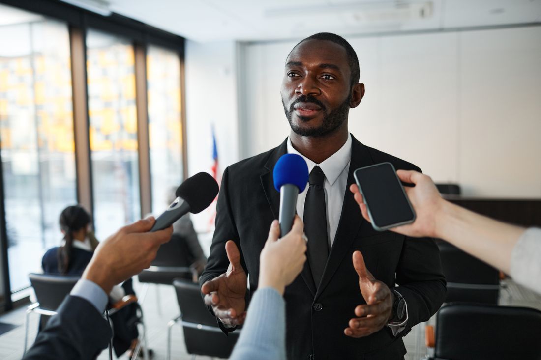 A businessman in a suit answers questions from several reporters holding microphones and a phone at a press conference.
