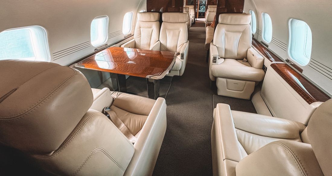 The empty cabin of a small private jet, cream leather seats facing a polished wood table between rows of oval windows.