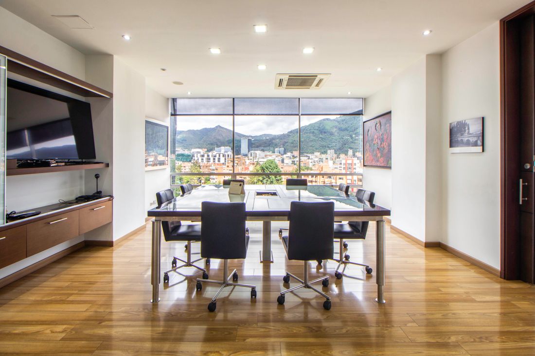 An empty high-floor boardroom with a long table, black leather rolling chairs, and floor-to-ceiling windows showing a city skyline and hills beyond.