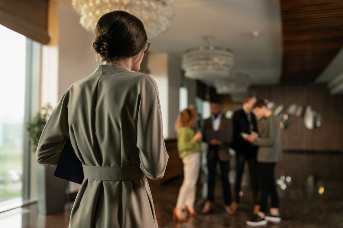 A person in a coat stands in the foreground of an elegant chandeliered foyer while a small, out-of-focus cluster of people in business attire talks in the background.