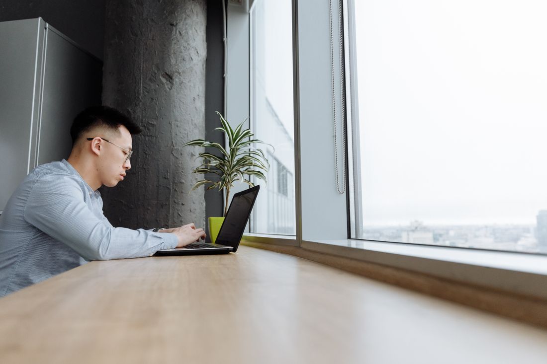 A man works quietly on a laptop at a window-side desk in a modern high-rise office, city view in the background.