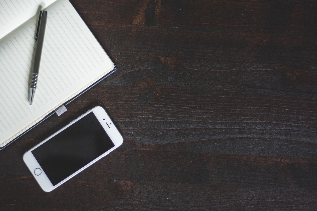 An open notebook with blank pages, a pen, and a phone with a dark screen sit together on a wooden desk.