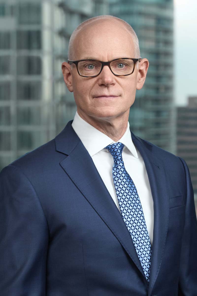 Bald man in glasses and a navy suit sharply in focus in front of a blurred office window and city skyline, showing a fast lens separating a subject from a busy background