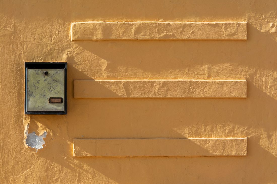 A single small metal mailbox on a plain yellow wall in hard afternoon shadow