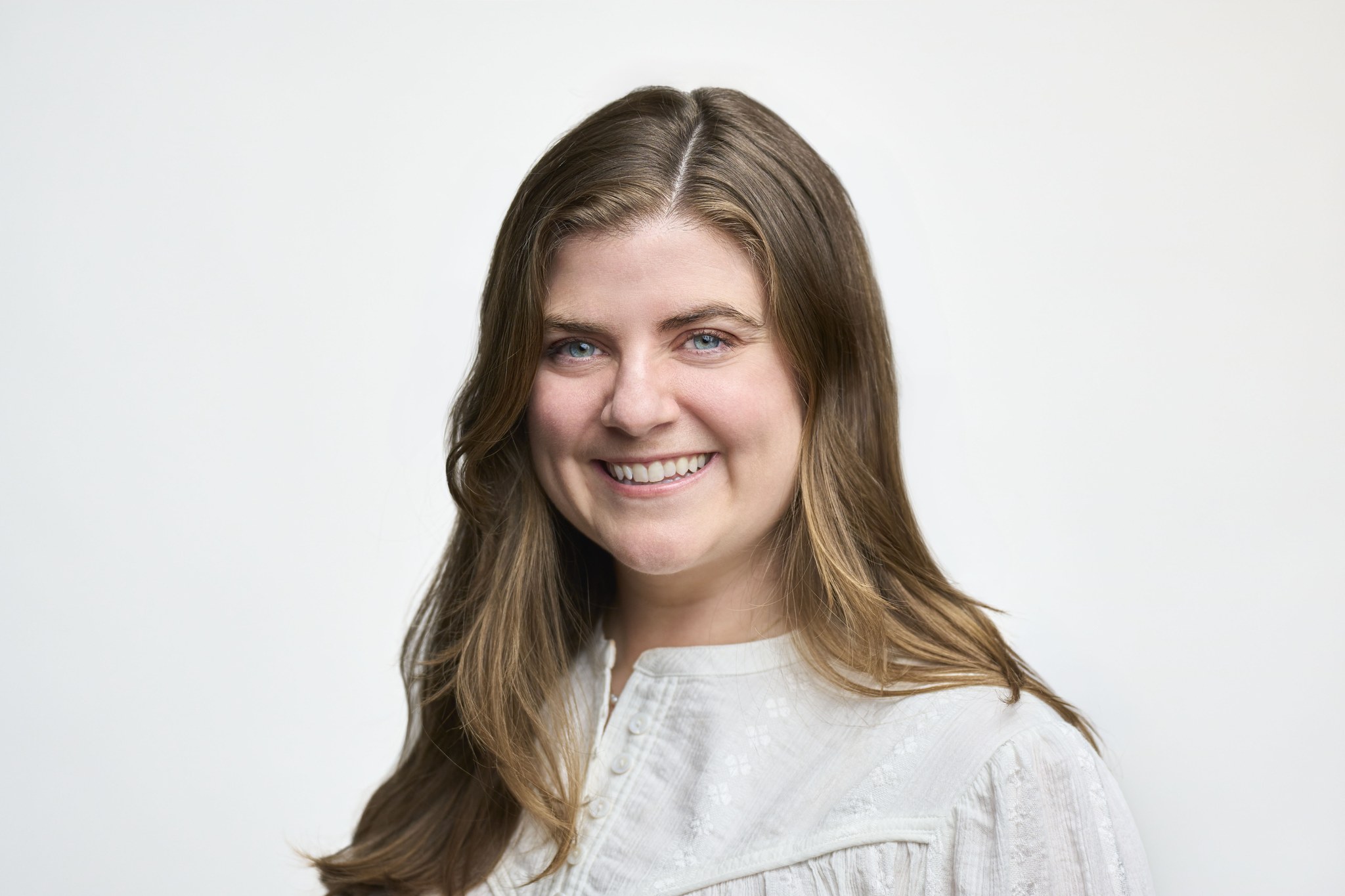 Studio headshot of a smiling woman with long brown hair wearing a white embroidered blouse.
