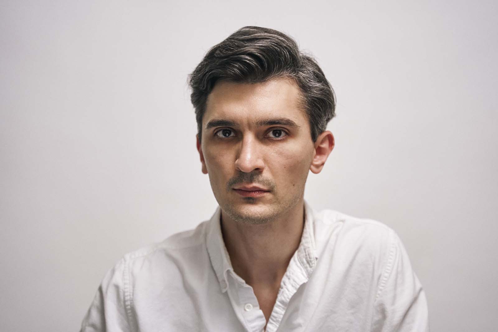 Close portrait of a man with wavy dark hair in a plain white shirt against a light background