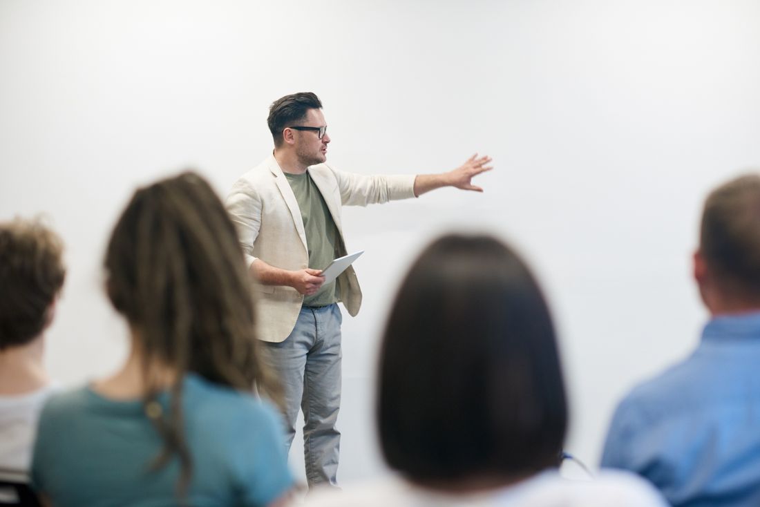 A man in a light blazer stands before a plain white wall, one arm extended mid-gesture, with blurred audience members in the foreground.